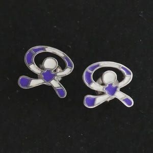 Könnte beinhalten: Ein Paar silberfarbene Ohrstecker. Jeder Ohrstecker zeigt eine stilisierte Figur mit weißem Zentrum, lila Umriss und lila Akzenten. Die Ohrringe sind vor einem dunklen Hintergrund platziert.