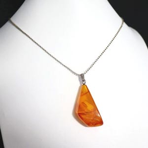 Puede incluir: Collar plateado con colgante de ámbar. El colgante de ámbar es de color naranja cálido y tiene forma triangular. El collar se exhibe sobre un maniquí blanco.