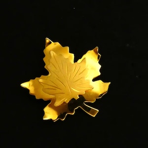 May include: A gold-coloured maple leaf brooch. The brooch is a detailed representation of a maple leaf, with intricate veining and a shiny, reflective surface. The brooch is set against a black background.