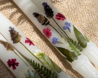 Op de afbeelding: Close-up van verschillende witte dinerkaarsen versierd met geperste bloemen en groen. De bloemstukken bevatten lavendel, rode en blauwe bloemen en groene bladeren. De kaarsen zijn gerangschikt op een getextureerd, beige oppervlak.