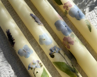 Puede incluir: Cuatro velas de marfil con flores y hojas prensadas. Las flores son azules, moradas y rosas. Las hojas son verdes.