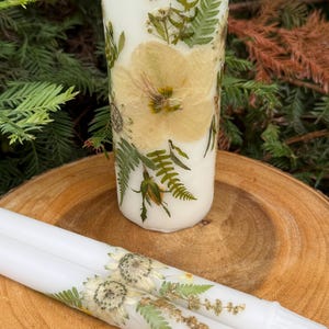 May include: A cylindrical white candle decorated with pressed yellow flowers and green fern leaves, accompanied by two slender white taper candles featuring similar pressed floral and fern botanical arrangements on their surfaces.