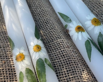 Puede incluir: Cuatro velas blancas en forma de pilar con margaritas blancas prensadas y hojas verdes. Las velas están dispuestas sobre una superficie de arpillera marrón.