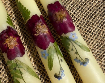 Op de afbeelding: Close-up van verschillende crèmekleurige taps toelopende kaarsen versierd met geperste bloemen. Elke kaars heeft een grote bordeauxrode bloem, kleine blauwe bloemen en groene bladeren. De kaarsen zijn gerangschikt op een getextureerd, bruin oppervlak.