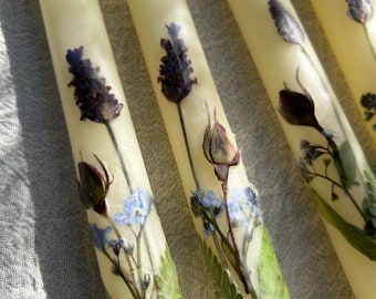 Op de afbeelding: Vier ivoorwitte kaarsen met geperste bloemen en bladeren die in de was zijn ingebed. De bloemen omvatten lavendel, vergeet-me-nietjes en rozen.