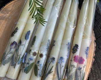 Op de afbeelding: Zes lange, witte kaarsen met geperste bloemen en groen. De kaarsen zijn verpakt in doorzichtig plastic en vastgebonden met touw.