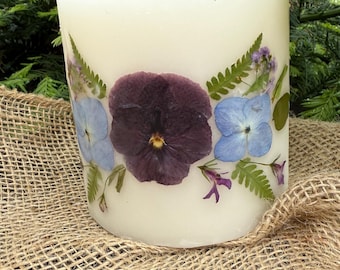 Puede incluir: Una vela blanca en forma de pilar con flores moradas y azules prensadas incrustadas en la cera. La vela está sobre una superficie de arpillera.