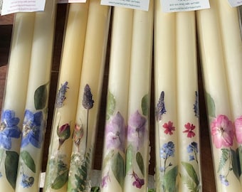 Op de afbeelding: Twee sets kaarsen met geperste bloemen en groen. De kaarsen zijn verpakt in doorzichtig plastic en hebben een label met de tekst "Wolf at the Door".