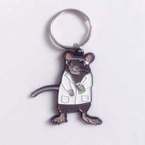 May include: A novelty keyring featuring a cartoon rat in a lab coat and safety glasses, holding a green beaker. The rat is brown and grey, with a silver key ring attached.