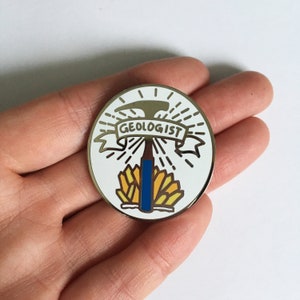 Puede incluir: Un pin de esmalte blanco con un contorno dorado. El pin presenta un pico con un mango azul, rodeado de cristales amarillos y una pancarta que dice "GEOLOGIST".