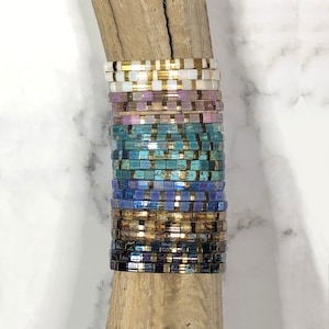 May include: A stack of colorful beaded bracelets with gold accents. The bracelets are arranged in a gradient pattern, starting with white and ending with dark blue. The bracelets are stacked on a piece of driftwood.