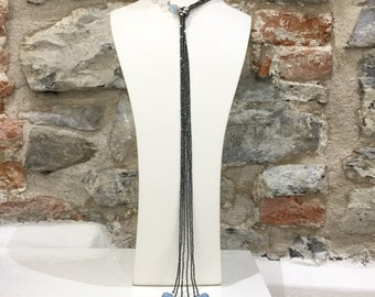 May include: A long lariat necklace featuring multiple thin, dark metallic chains that drape downward. The necklace is accented with light blue spherical beads at the ends and near the neck, complemented by small silver-toned decorative spacers.