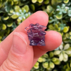 May include: A small purple fluorite crystal carved into the shape of a cartoon character. The crystal is translucent and has a slight shimmer.