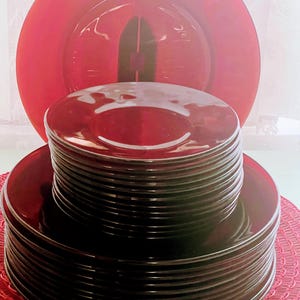 Puede incluir: Una pila de platos de vidrio rojo oscuro, con un gran plato de carga rojo en el fondo. Los platos están colocados sobre un salvamanteles rojo, creando un juego de comedor cohesivo. Los platos son de un color rojo intenso y rico.