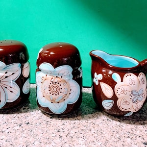 May include: A set of three brown ceramic kitchenware pieces with a floral design in shades of blue, white, and brown. The set includes a creamer, a salt shaker, and a pepper shaker.