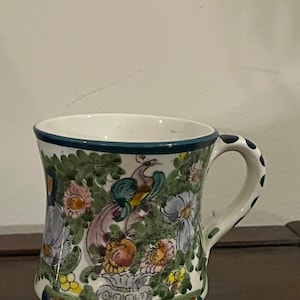 May include: A white ceramic mug with a blue rim and handle. The mug is decorated with a floral pattern in shades of green, pink, yellow, and blue. The flowers are surrounded by green leaves and a small bird.