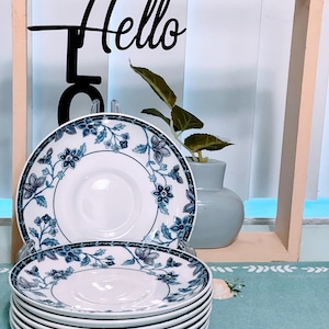 May include: A stack of white ceramic plates with a blue floral design. The plates are stacked in front of a decorative wooden frame with the word "Hello" in black script. A small vase with a plant is visible in the background.