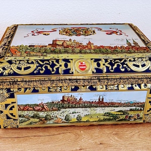 May include: A vintage tin box with a gold and blue design. The lid features a detailed illustration of a city skyline with a red and white logo that says "Schmidts". The bottom of the box shows a similar illustration of the city with a river running through it.