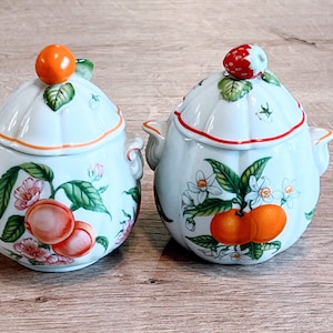 May include: Two white ceramic jars with lids. One jar features a peach design, the other an orange design. Both jars have handles and are decorated with fruit and floral patterns. The lids are topped with a fruit-shaped knob.