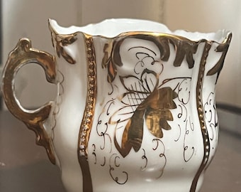 Puede incluir: Taza de té de porcelana blanca con detalles dorados. La taza tiene un asa dorada y está adornada con patrones y adornos florales dorados. El borde de la taza tiene un borde festoneado. La taza de té es probablemente vintage o antigua.