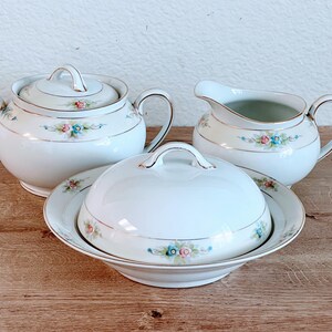 Puede incluir: Un azucarero de porcelana blanca con tapa, una jarra de crema y una mantequillera con tapa. Las tres piezas tienen un diseño floral con flores rosas, azules y verdes.