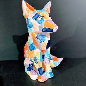 Puede incluir: Una figurita de zorro de cerámica colorida con un patrón geométrico en tonos de azul, naranja, amarillo, rosa y gris. El zorro está sentado con la cola enroscada alrededor de sus patas.