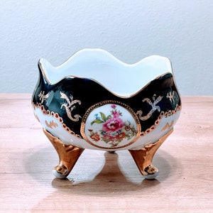May include: A white porcelain bowl with a scalloped rim and three gold-painted feet. The bowl is decorated with a black and gold floral design and a central floral motif.