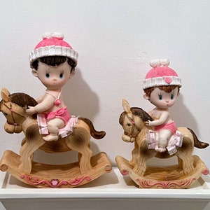 May include: Two ceramic figurines of babies riding rocking horses. The babies are wearing pink and white hats and dresses. The rocking horses are brown and white.