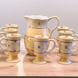 Puede incluir: Un juego de tazas de cerámica amarilla y marrón con diseños florales y una jarra a juego. Las tazas tienen una base con pie y son perfectas para servir café o té.