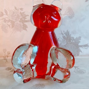 May include: A red glass bear figurine with clear glass arms and legs. The bear is sitting on a white surface.