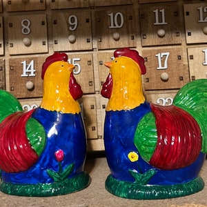 Könnte beinhalten: Zwei Keramik-Salz- und Pfefferstreuer in Form von Hähnen. Die Hähne sind in leuchtenden Farben bemalt, darunter Rot, Gelb, Blau und Grün. Sie stehen sich gegenüber und haben einen verspielten, volkstümlichen Stil.