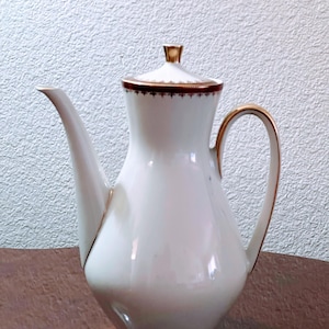 Puede incluir: Tetera de porcelana blanca con ribete dorado y tapa. La tetera tiene una base ancha y un pico estrecho.