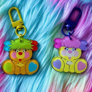 May include: Two colorful keychains featuring cartoon bear designs. The bears are yellow with green, red, and blue accents. Each keychain has a metal clasp in yellow or blue. The background is a vibrant, multicolored faux fur.