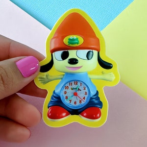 May include: A yellow sticker featuring a cartoon dog wearing a red hat with a green frog on it. The dog is holding a blue clock with a red hand pointing to the number 12. The clock face has the numbers 1 through 12 and the words "Snooze" and "Alarm".
