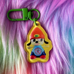May include: A yellow keychain featuring a cartoon character wearing an orange beanie with a small green frog emblem. The character has a light blue shirt with an analog clock face on the chest, dark blue pants, and red shoes, attached to a green clip.