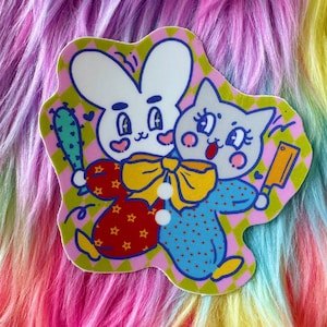 May include: A colorful sticker featuring a cartoon rabbit and cat embracing. The rabbit wears a red outfit with star accents and holds a cactus club. The cat is in a blue polka-dot outfit and holds a cleaver. The background is a pink and green checkered pattern.
