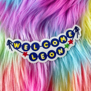 May include: A sticker featuring a string of blue circular beads with yellow letters spelling out WELCOME LEON. The design includes small red and green star accents at the ends and between the words, all set against a white die-cut border.