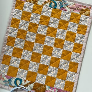 Könnte beinhalten: Handgefertigter, gesteppter Tischset mit einem Schachbrettmuster aus senfgelben und grauen Quadraten. Der Rand umfasst Stoff mit Blumen- und geometrischen Mustern in Rosa, Gold und Blau. Unten ist ein hellblaues Band befestigt.