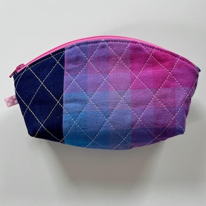 Peut inclure: Une trousse de maquillage matelassée avec un dégradé de couleurs allant du bleu marine au violet et au rose. Le sac a une fermeture éclair rose et une petite boucle en tissu sur le côté. Le tissu présente un motif en losange créé par des coutures blanches.