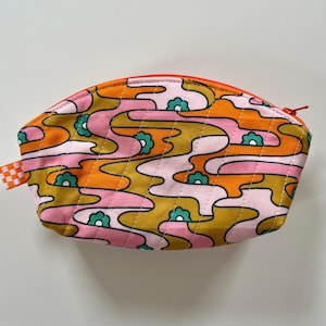 May include: A small, quilted cosmetic bag with a vibrant, retro-inspired pattern. The design features flowing, abstract shapes in pink, orange, and gold, accented with teal flower motifs. The bag has a red zip and a small checkered fabric loop.
