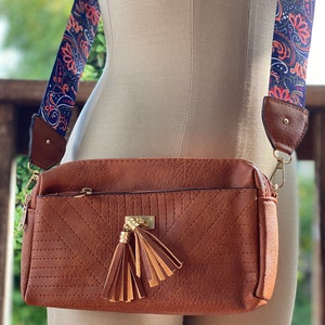 May include: A brown faux leather crossbody bag with a gold zipper and a tassel. The bag has a patterned strap with blue, orange, and green colors.