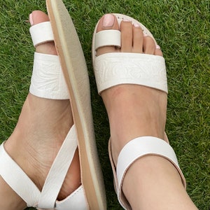 May include: White leather sandals with a toe ring and ankle strap. The sandals have an embossed floral design and a light brown sole.
