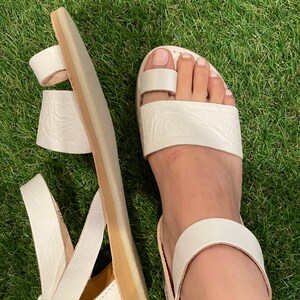 May include: White leather sandals with a strap around the ankle and a toe loop. The sandals have a textured design and a light brown sole.