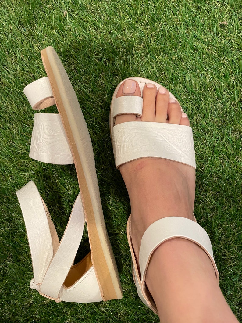 May include: White leather sandals with a strap around the ankle and a toe loop. The sandals have a textured design and a light brown sole.