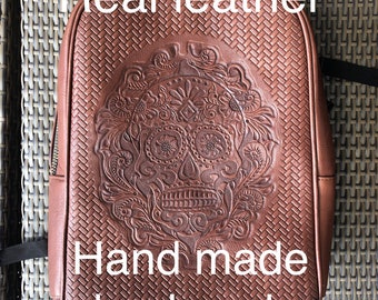May include: A brown leather backpack with a tooled design of a sugar skull. The backpack has the text "Real leather" and "Hand made backpack" on it.