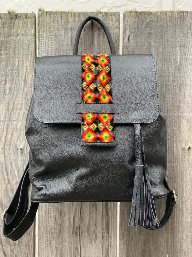 Op de afbeelding: Zwarte leren rugzak met een kleurrijke kralenband. De band heeft een geometrisch patroon in tinten rood, oranje, geel en groen.