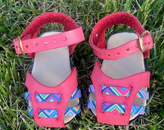 May include: A pair of red leather sandals with colorful patterned straps. The sandals have an adjustable ankle strap with a gold buckle.