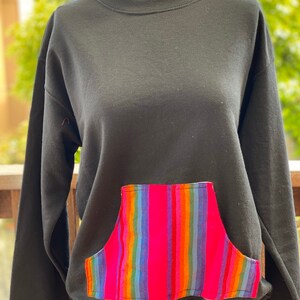 May include: Black sweatshirt with a colorful striped pocket. The pocket is made of a woven fabric with pink, blue, green, orange, and yellow stripes.