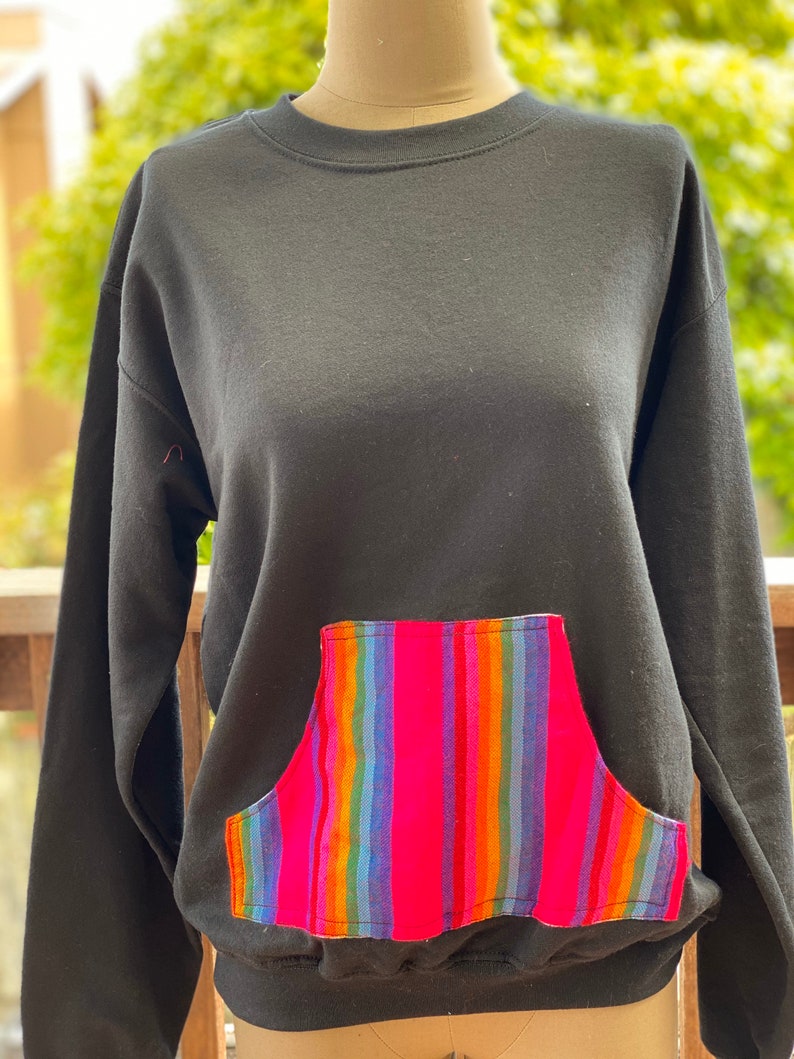 May include: Black sweatshirt with a colorful striped pocket. The pocket is made of a woven fabric with pink, blue, green, orange, and yellow stripes.
