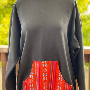 May include: Black sweatshirt with a large, colorful, patterned pocket on the front. The pocket has a geometric design with red, pink, blue, and white stripes.